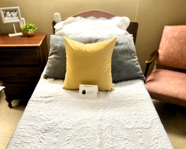 A neatly made single bed with a white quilted bedspread, a yellow pillow, and two gray pillows. A small card with the Amistad Nursing & Rehabilitation Center logo is placed on the bed. To the left of the bed is a wooden nightstand with a small green plant and a framed photo. To the right is a pink upholstered armchair.