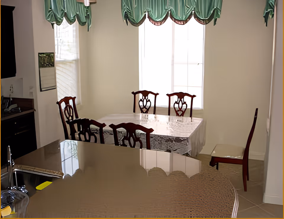 Dining room with a table covered by a tablecloth surrounded by wooden chairs, green valance curtains over two windows, and a kitchen counter in the foreground.