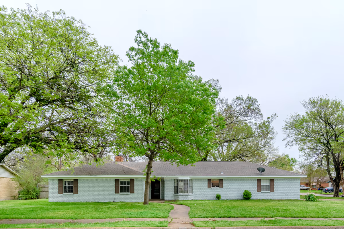 Front view of a single-story blue brick home with a green lawn and large trees.