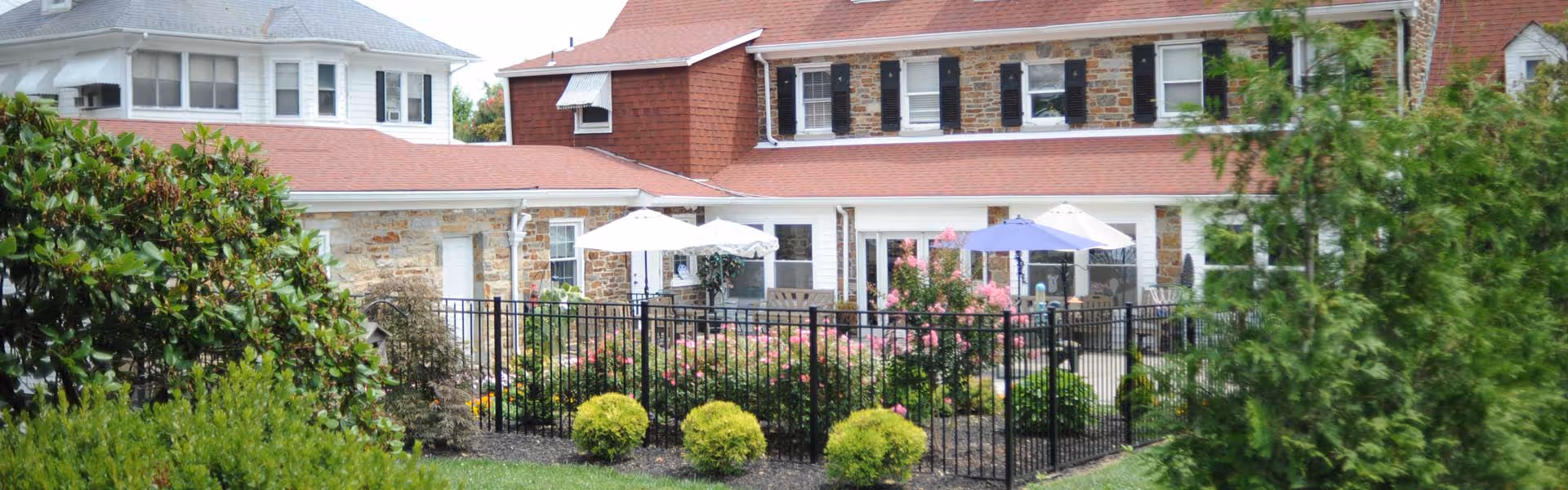 Outdoor view of a senior living facility with a stone and brick building featuring multiple windows and red roofs. In front of the building, there is a fenced garden area with green shrubs, flowering plants, and patio umbrellas providing shade over outdoor seating.