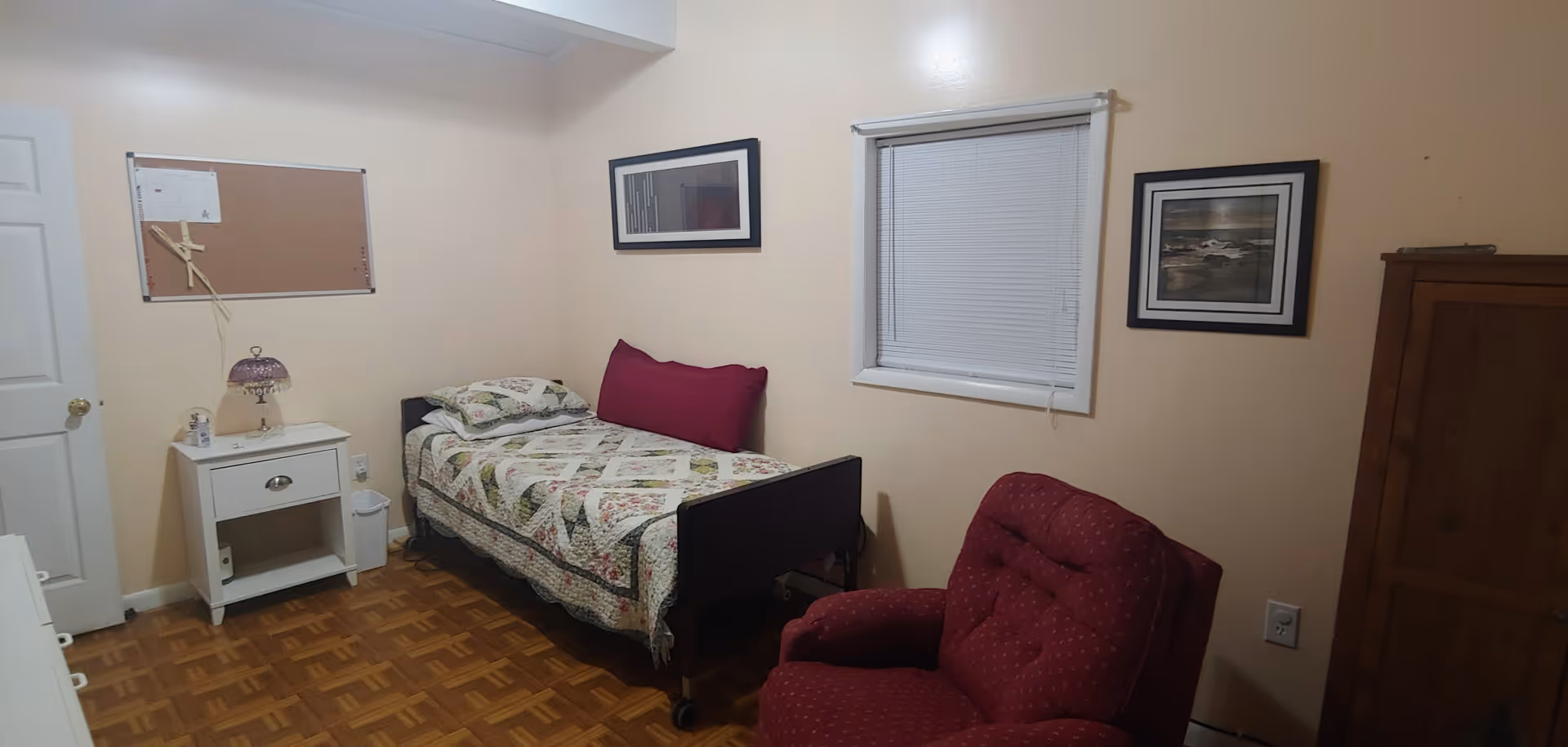 A small bedroom with a single bed covered in a floral quilt and a large red pillow. Next to the bed is a white nightstand with a decorative lamp and a small trash bin underneath. There is a red upholstered armchair in the foreground, a window with closed blinds on the wall, and two framed pictures hanging on the walls. The floor is wooden with a parquet pattern.