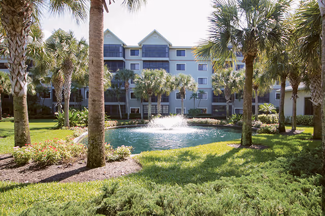 A multi-story blue-gray senior living building surrounded by palm trees and a central fountain pond.