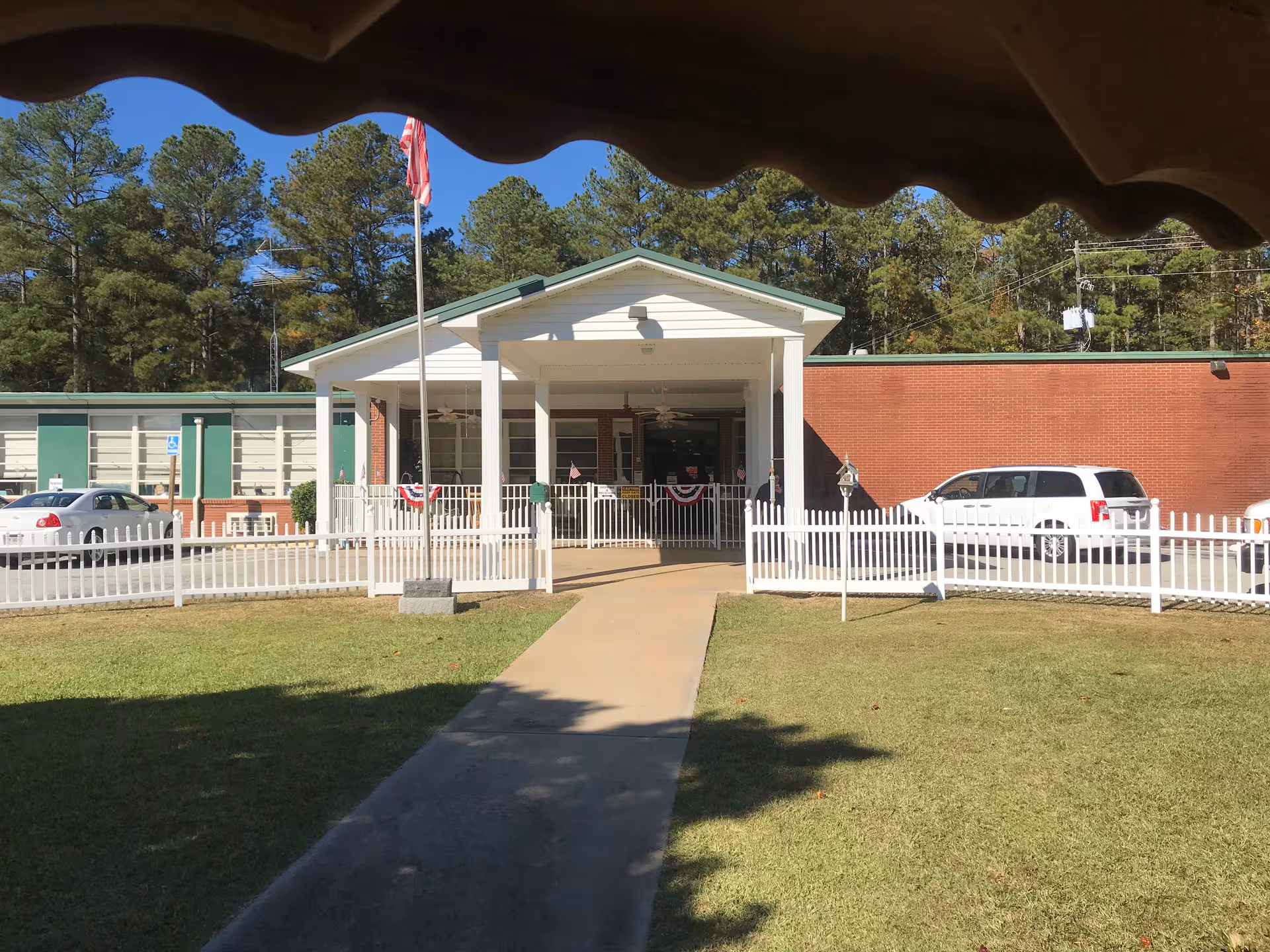 Exterior view of Heardmont Nursing Home entrance with a white fence, flag, and vehicles.