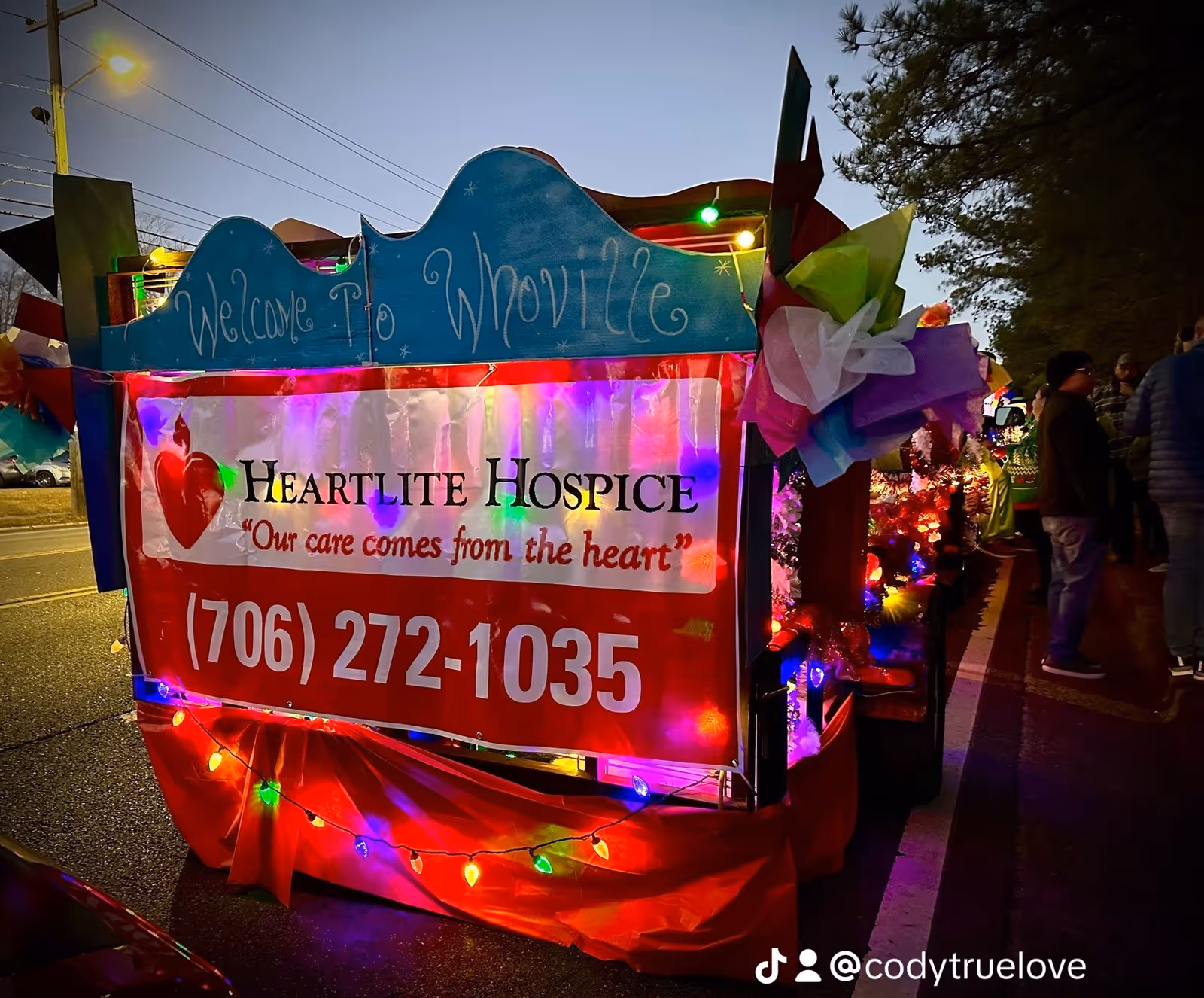Colorful parade float advertising Heartlite Hospice with string lights along a city street.