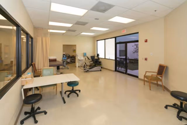Bright rehabilitation/therapy room with exercise equipment, a treatment table and privacy curtain, several stools and chairs, and glass doors to the outside.