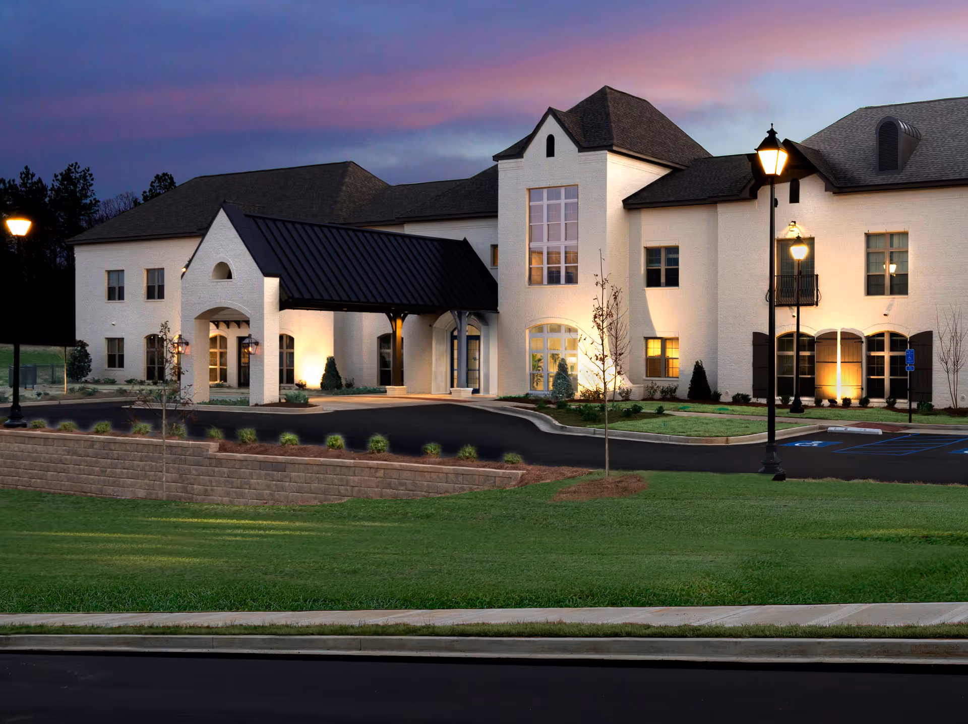 Exterior view of a large, white senior living facility building with a covered entrance, lit street lamps, and a well-maintained lawn at dusk with a purple and pink sky.