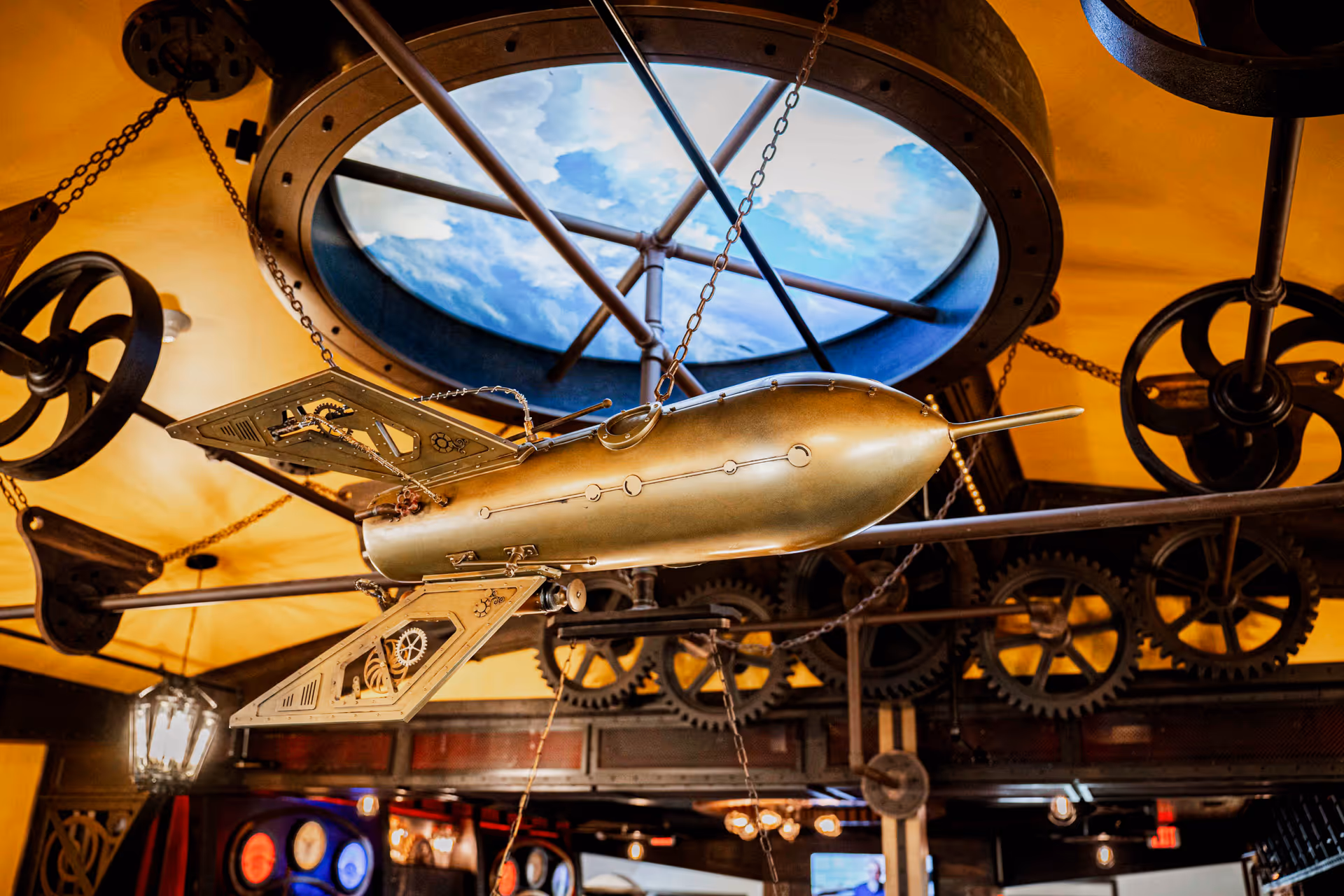 A steampunk-style interior ceiling decoration featuring a metallic, vintage submarine model suspended by chains. The ceiling has a circular skylight with a painted sky and clouds, surrounded by large gears and mechanical wheels. Warm lighting and industrial design elements are visible in the background.