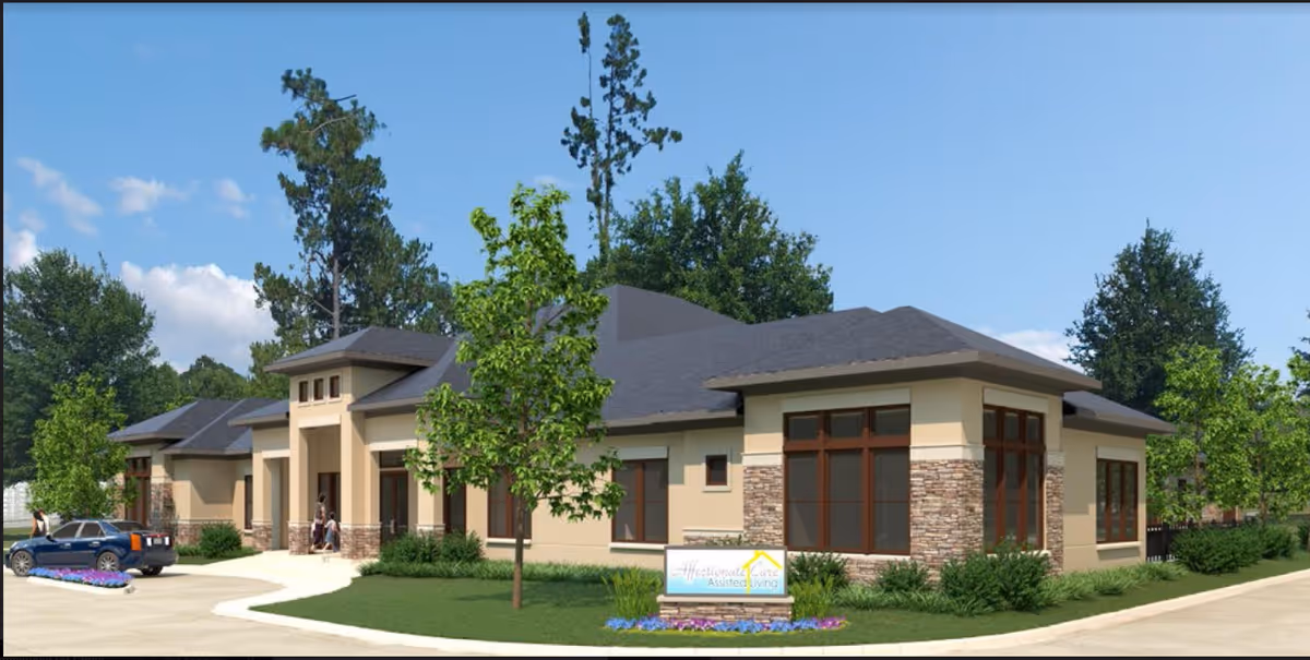 Exterior view of a single-story assisted living facility building with beige walls, stone accents, large windows, and a dark gray roof. There is a driveway with a parked blue car, a few people walking near the entrance, green trees, shrubs, and a sign in front that reads 'Affectionate Care Assisted Living'. The sky is clear and blue.