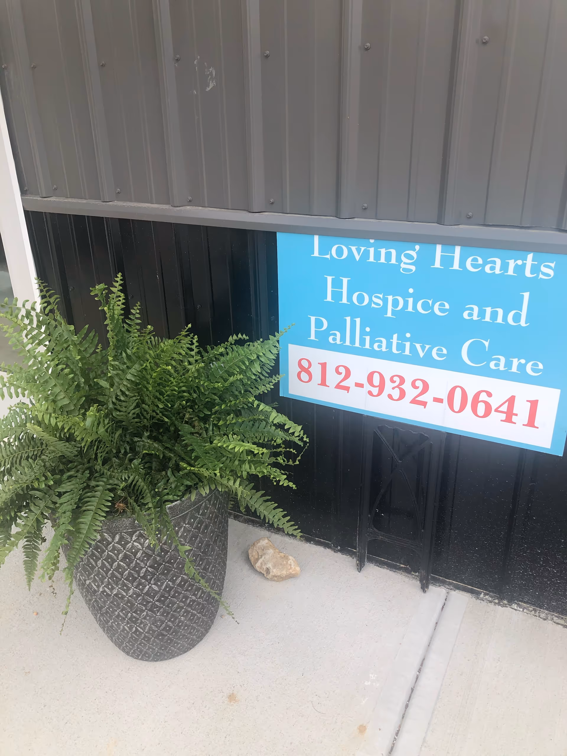 Exterior view of a building with a blue hospice sign and potted fern.