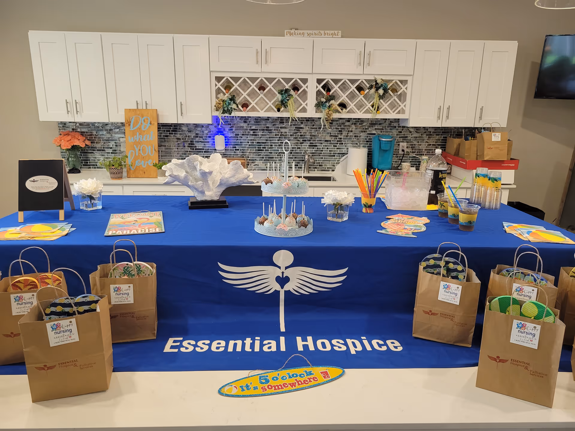 Blue tablecloth covers a table with Essential Hospice banner and gift bags in a kitchen backdrop.
