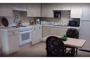 A compact apartment-style kitchen with white cabinets, stove, sink, fridge, and a small dining table.
