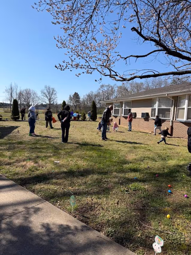Group of people outside a single-story brick facility on a sunny day in a grassy yard.