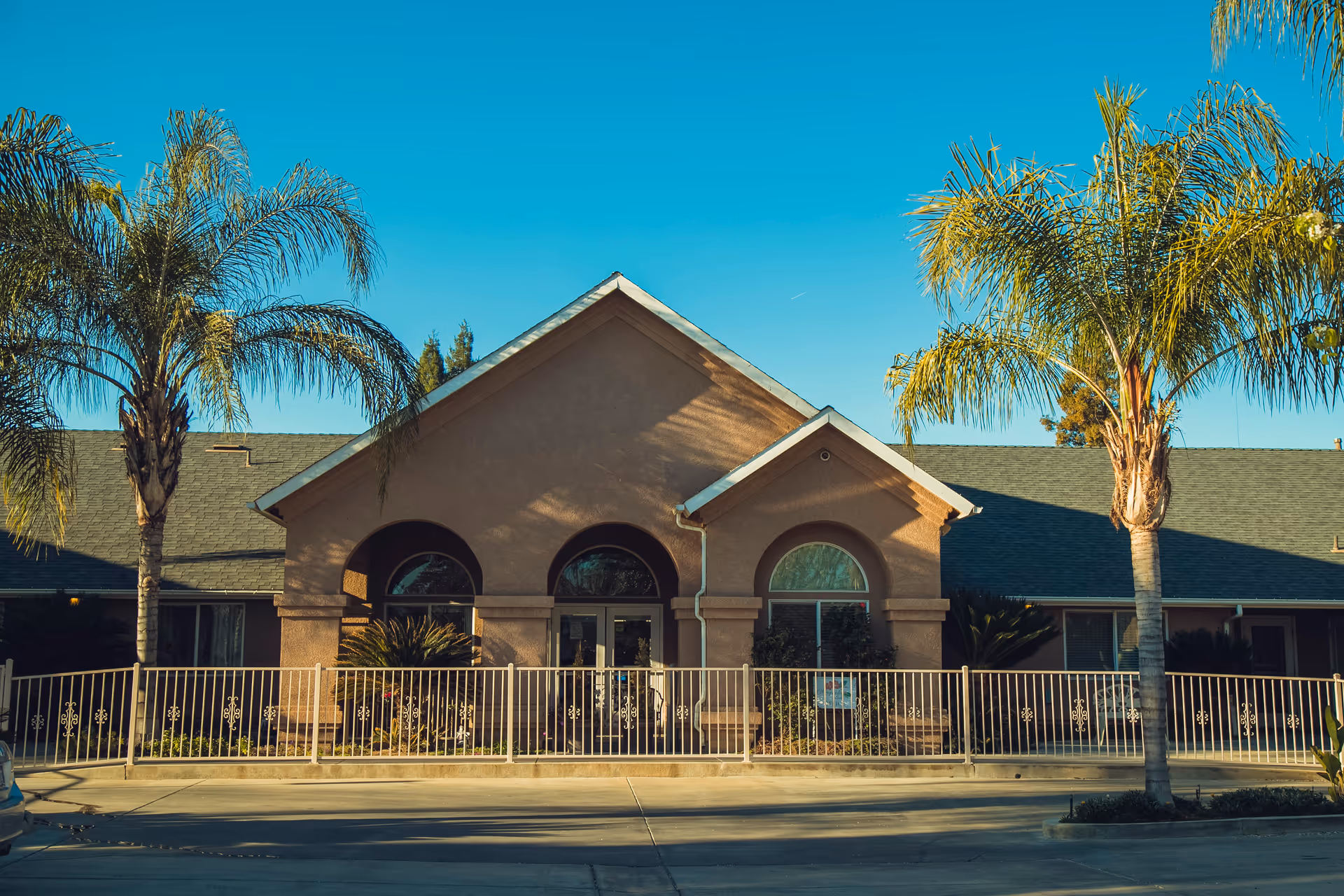 Front view of a senior living facility with arched windows, palms, and railing.