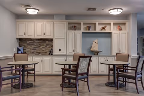 A bright senior living common area with white cabinetry, round tables, and chairs.
