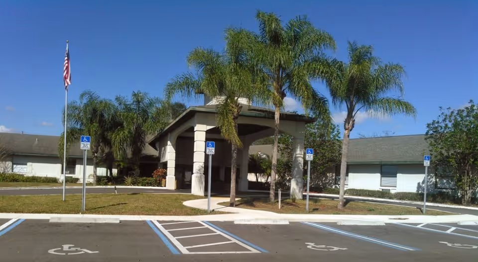 Exterior view of Sunny Hills Alf - Assisted Living Facility Sebring showing the entrance with a covered drop-off area, palm trees, an American flag, and several handicap parking spaces in front under a clear blue sky.