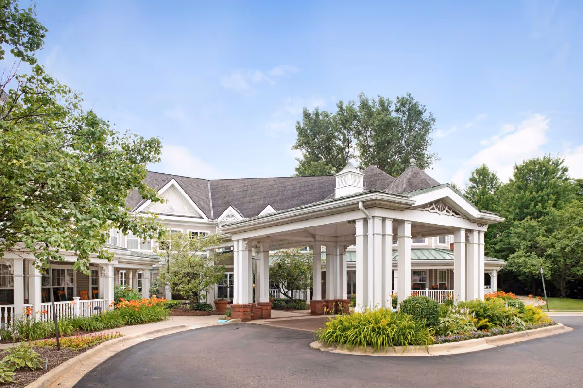 Exterior view of a senior living facility entrance with a covered drop-off area, surrounded by well-maintained landscaping including green shrubs, flowers, and trees under a partly cloudy sky.