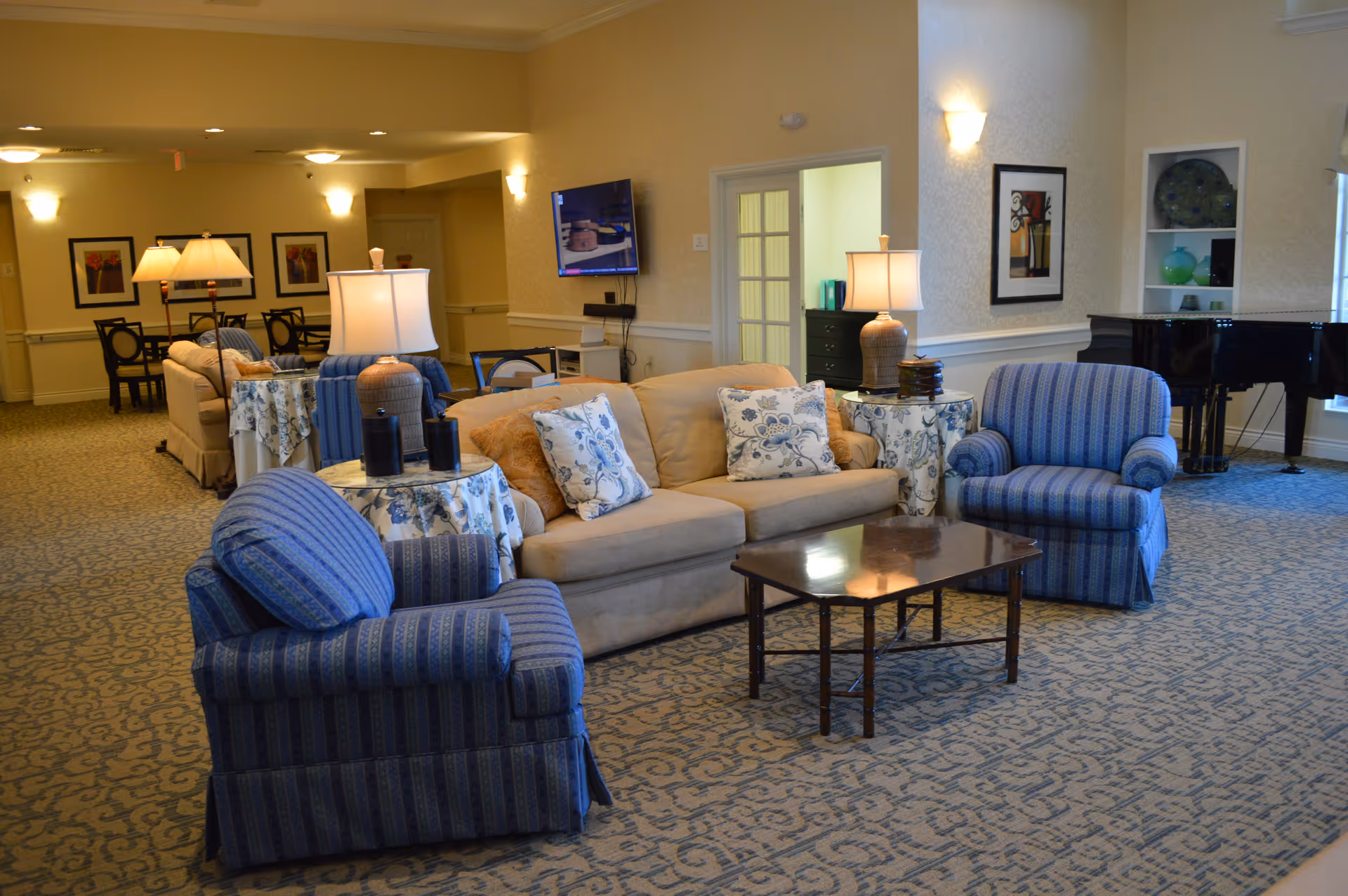 A cozy senior living facility common area with a beige sofa adorned with floral cushions, two blue striped armchairs, and a wooden coffee table. The room is softly lit with table lamps on side tables covered with floral cloths. In the background, there is a dining area with a table and chairs, framed artwork on the walls, a flat-screen TV mounted on the wall, and a black grand piano near a window.