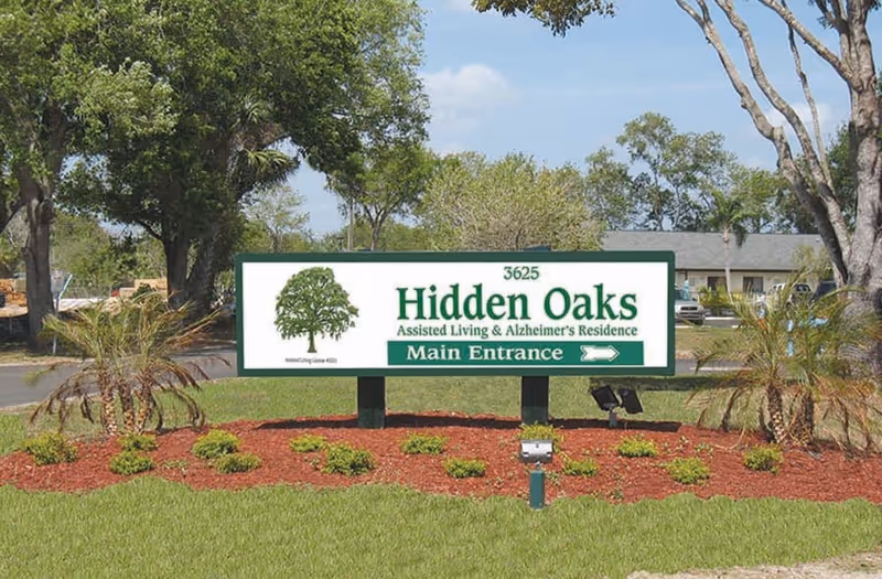 Outdoor view of a large green and white sign for Hidden Oaks Assisted Living & Alzheimer's Residence with the address 3625 and an arrow pointing to the main entrance, surrounded by landscaped grass, small plants, and trees under a clear sky.