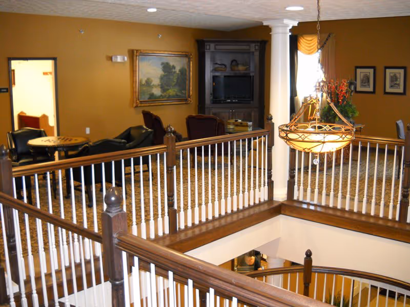 Interior view of a senior living facility lounge area with a wooden railing overlooking a lower floor. The room features a carpeted floor, a large hanging chandelier, a wooden entertainment center with a TV, several chairs, a small table, framed artwork on the walls, and a window with drapes.