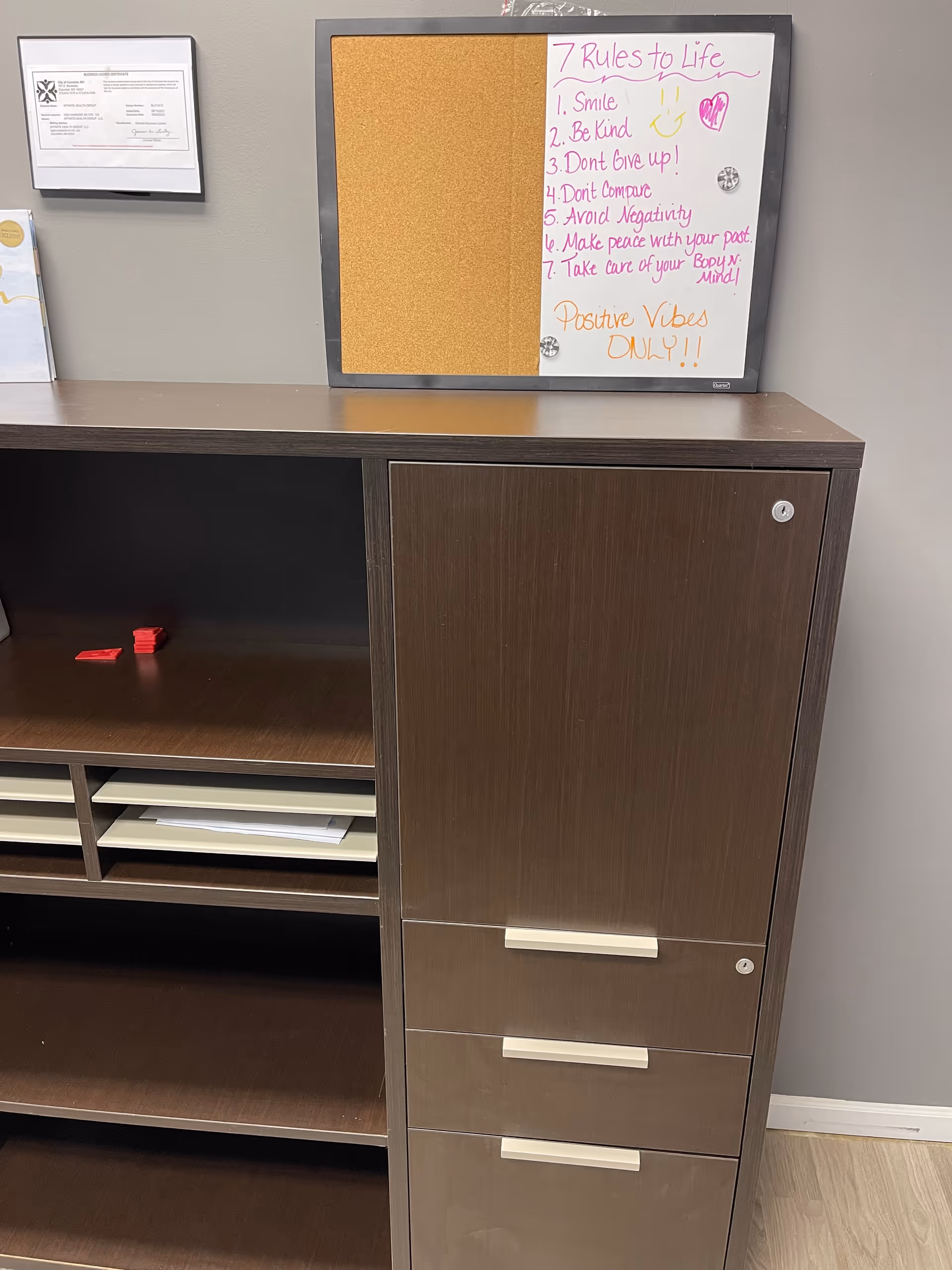 A dark brown modular cabinet and corkboard with handwritten rules sits against a gray wall.
