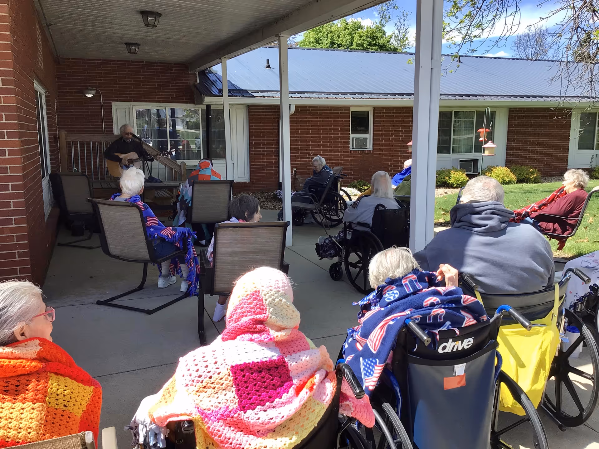 A group of elderly residents wrapped in colorful blankets sit in wheelchairs and chairs on a covered patio watching a musician play guitar.