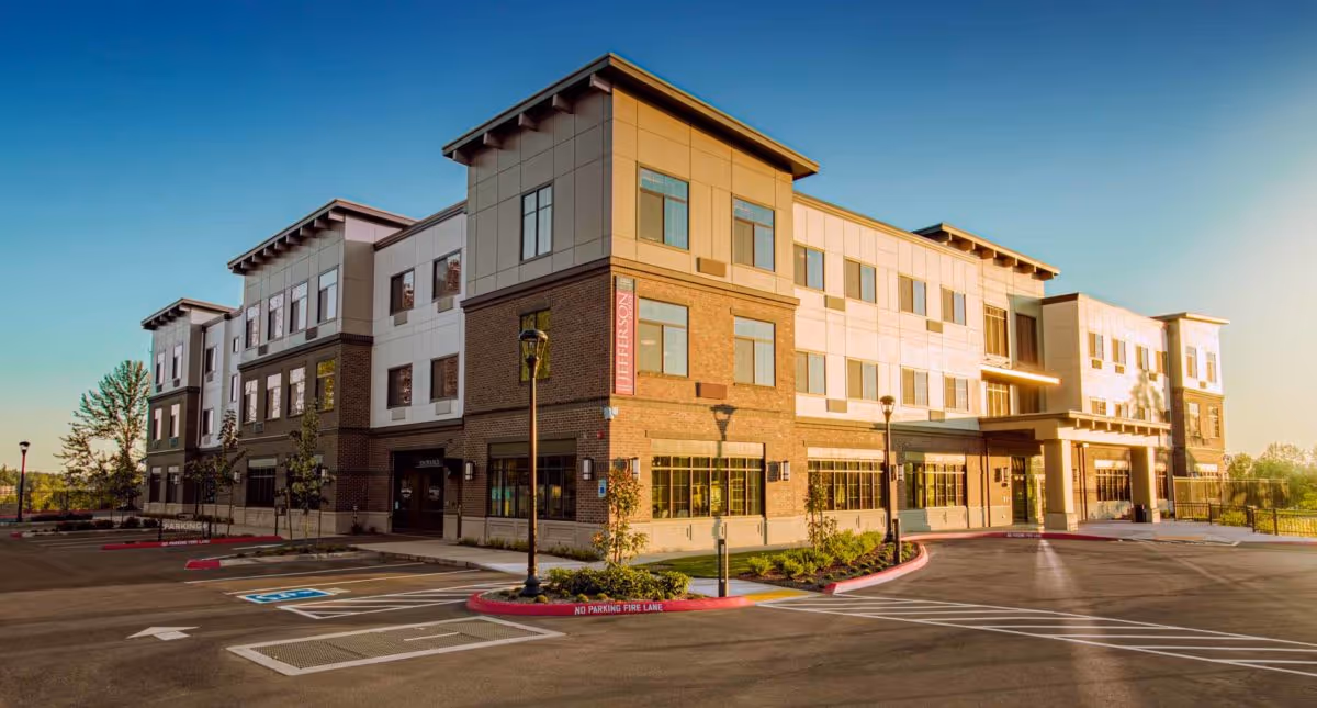 Front exterior of a modern three-story memory care building with a parking lot and landscaping at sunset.