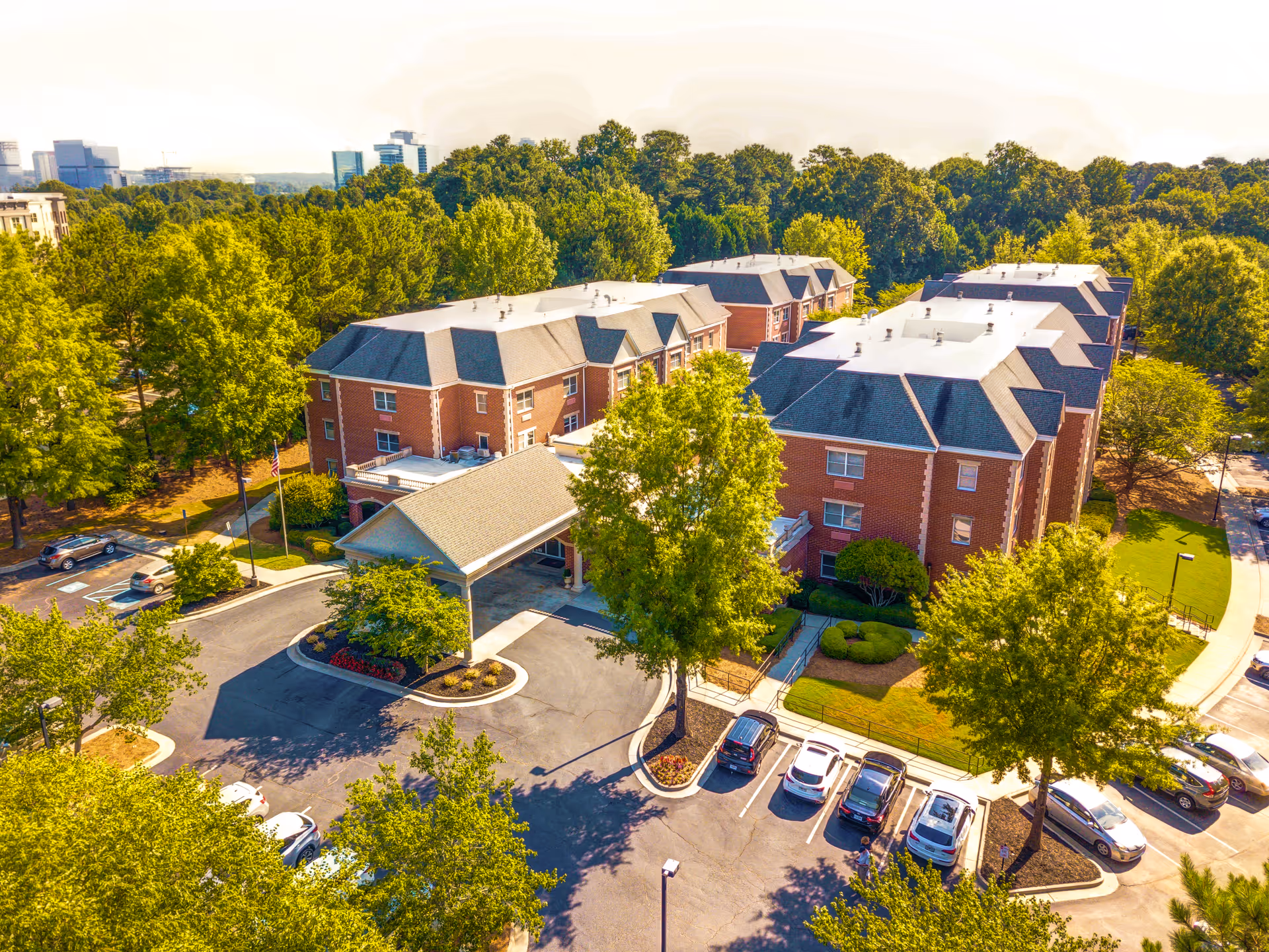 Aerial view of Dunwoody Place senior living facility showing multiple three-story brick buildings surrounded by trees and greenery. There is a covered entrance with a driveway and several parked cars in the parking lot. The background includes a distant city skyline.