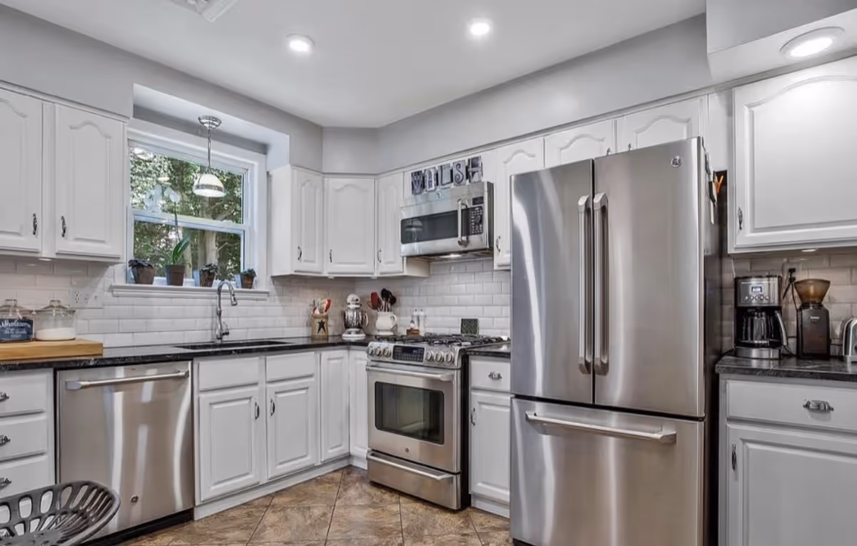 Bright white kitchen with stainless steel appliances, sink under window, tiled floor.
