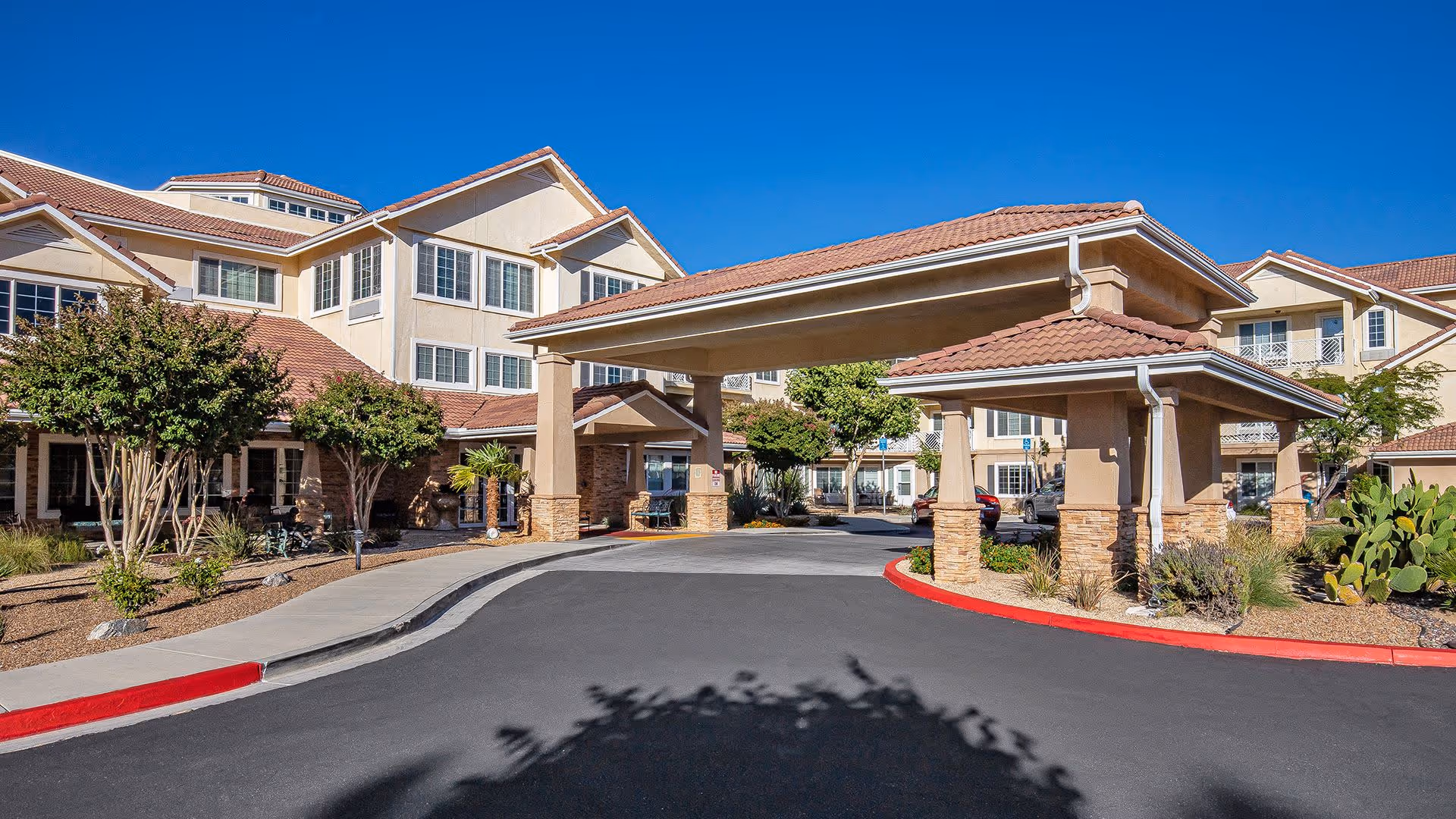 Exterior view of Holiday Rancho Village senior living facility showing a large covered entrance with beige pillars and stone accents, surrounded by a well-maintained driveway, landscaping with trees and shrubs, and a clear blue sky.