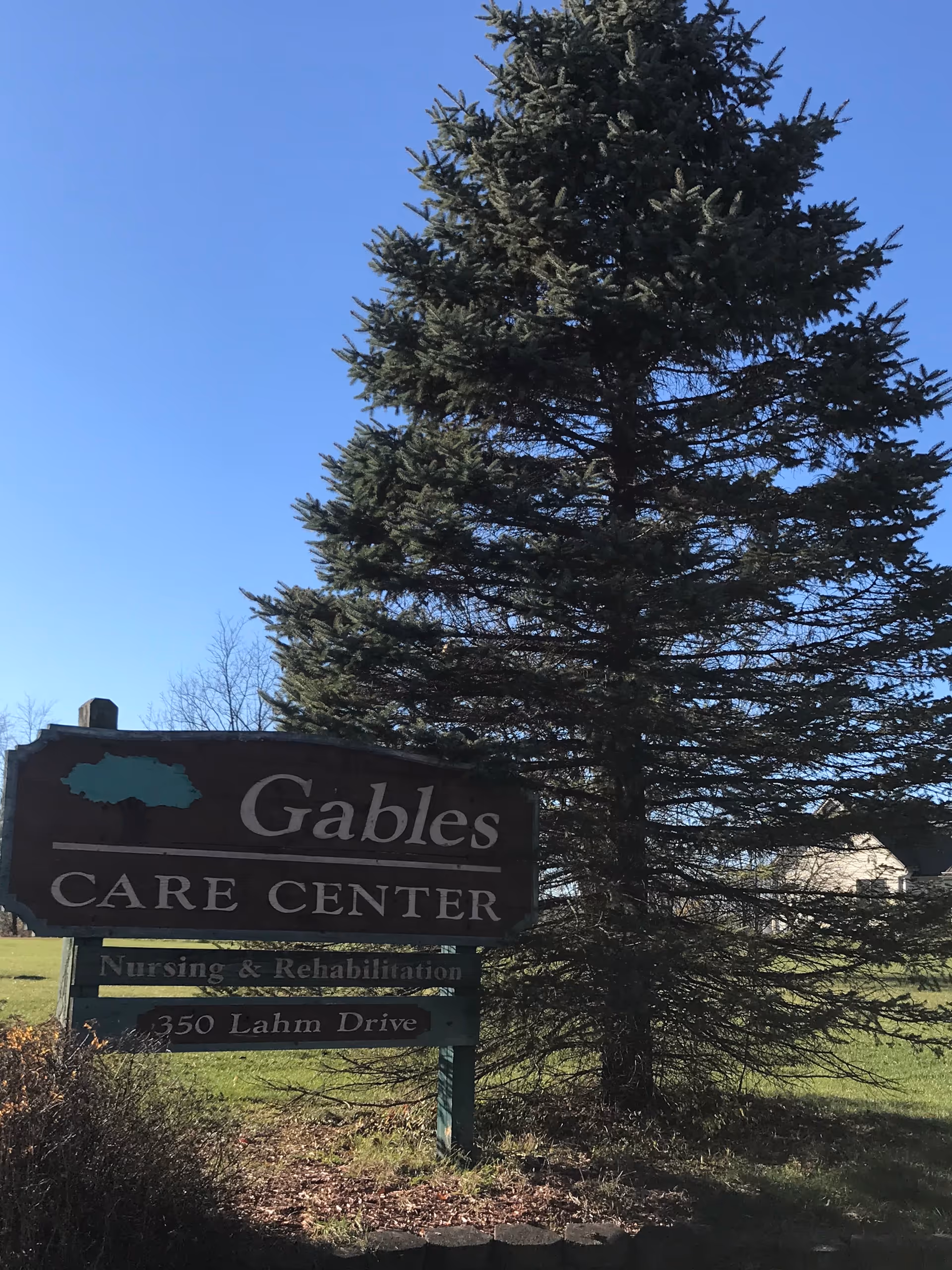 Sign for Gables Care Center on a grassy lawn with a tall evergreen tree.