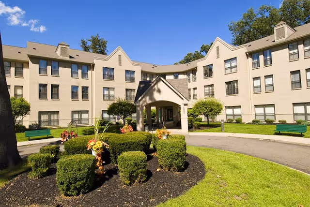 Exterior view of McGivney-Bethune Apartments, a three-story beige building with multiple windows. The foreground features a circular driveway with landscaped bushes and flower arrangements, green benches, and a covered entrance. The sky is clear with a few clouds.