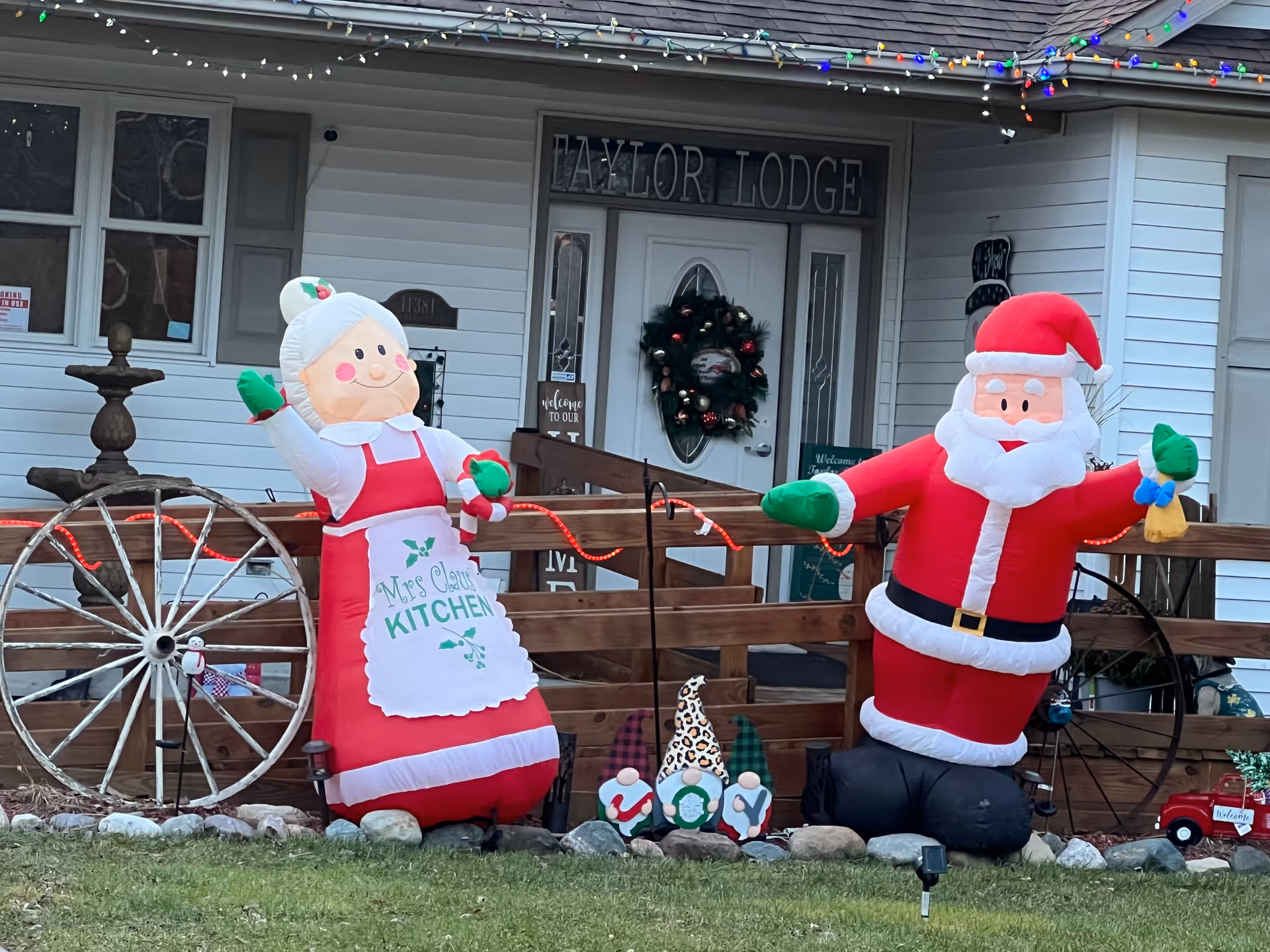 Front entrance of Taylor Lodge decorated for Christmas with inflatable figures of Mrs. Claus and Santa Claus on either side of the walkway. There is a wooden fence with a wagon wheel, a Christmas wreath on the door, and holiday lights hanging from the roof.