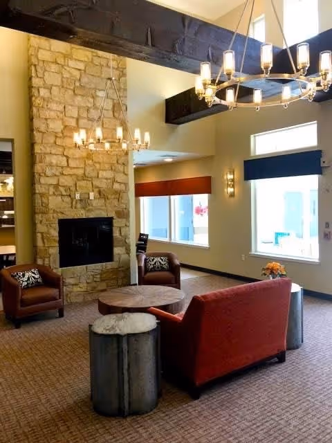 A cozy living room area with a stone fireplace, two brown armchairs, a red sofa, a round wooden coffee table, and two cylindrical side tables. The room features high ceilings with exposed dark wooden beams and two large chandeliers. There are large windows with blue and red valances letting in natural light.