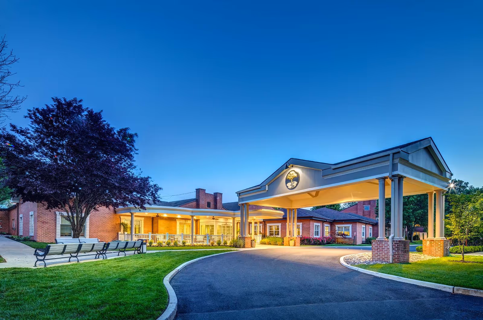 Front entrance of a brick senior living facility at dusk with a covered porte-cochere, benches, and illuminated walkway.