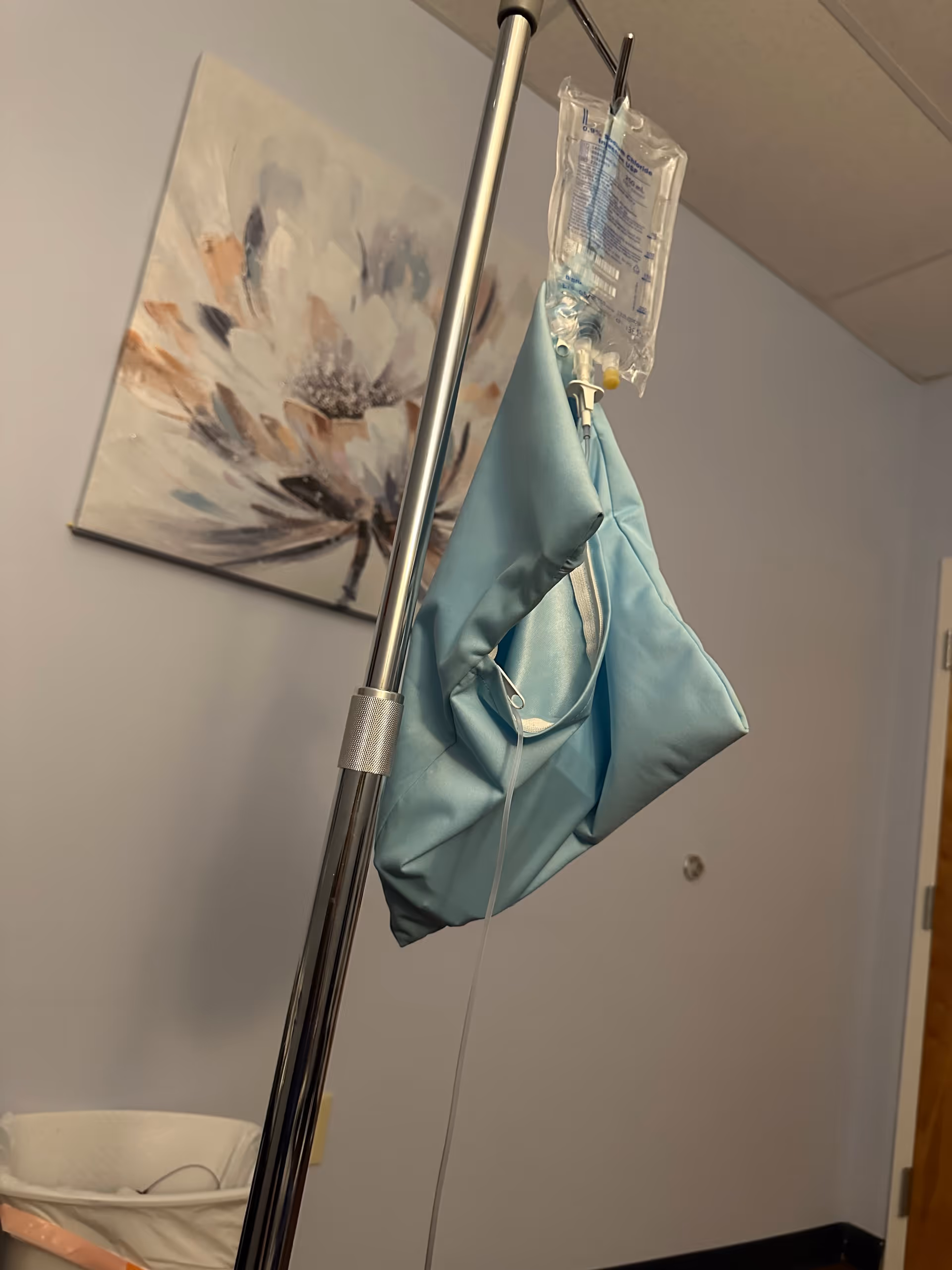 Medical IV pole with bag hangs beside a blue clinical bag in a hospital-style room.