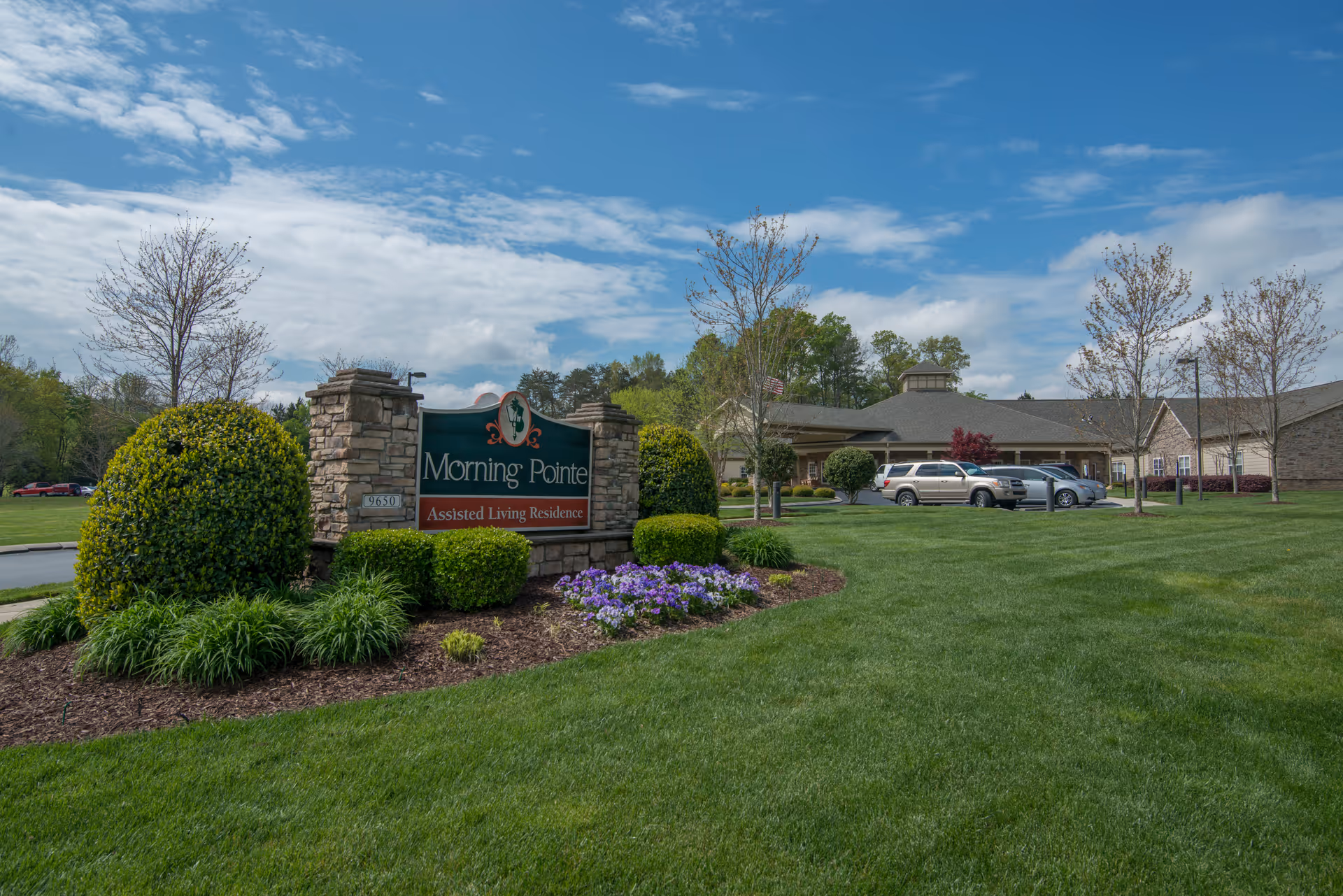 Exterior view of Morning Pointe of Greenbriar Cove assisted living residence with a stone sign surrounded by manicured bushes and flowers, a green lawn, several trees, parked cars, and a partly cloudy blue sky.
