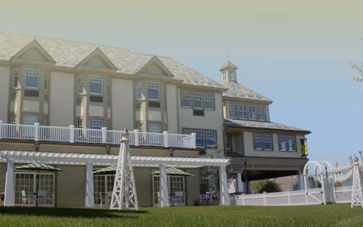 Exterior view of a multi-story senior living facility with beige siding, multiple windows, and a white railing balcony. The building has a small tower-like structure on the roof and a green lawn in the foreground with white decorative structures and a white fence.