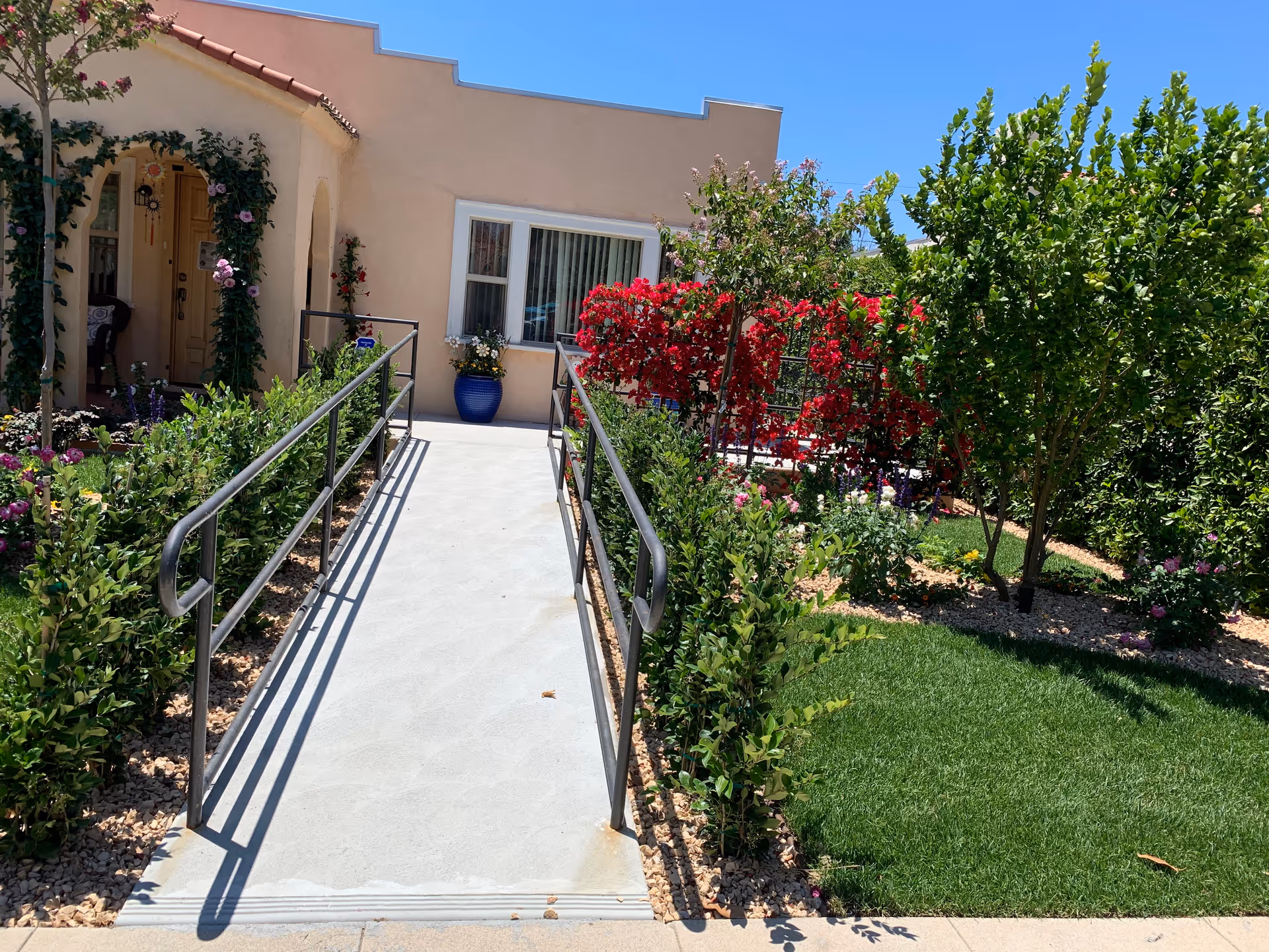 Ramp leading to a beige senior living facility entrance with a colorful garden and shrubs.