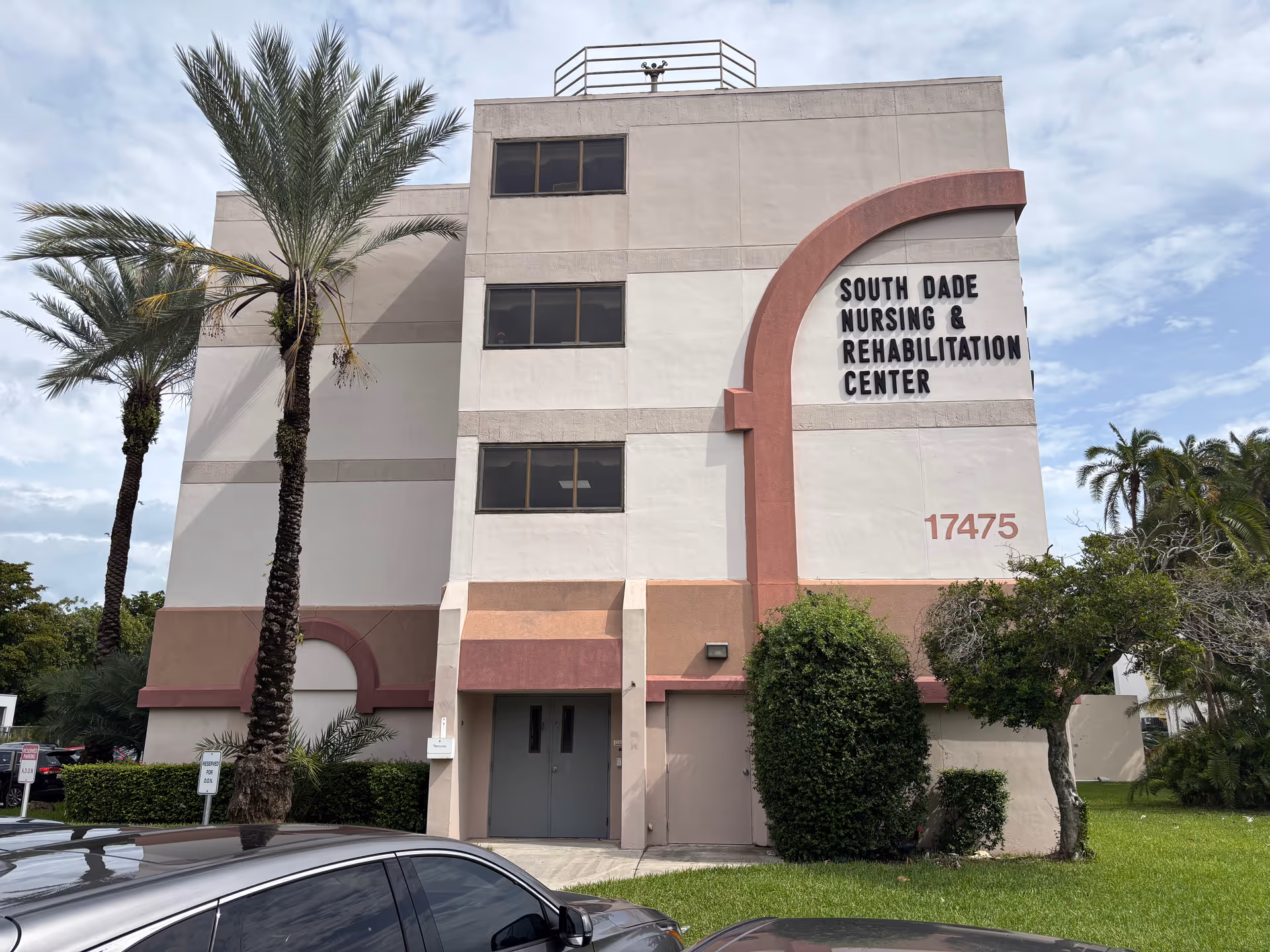 Exterior view of South Dade Nursing & Rehabilitation Center building with palm trees and cars.