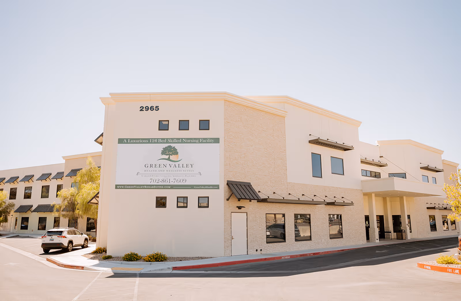 Exterior view of Green Valley Health & Rehabilitation, a two-story building with beige and white walls, multiple windows, and a large sign displaying the facility's name, contact number, and website. There are a few cars parked near the building and some small trees and shrubs around the parking area.