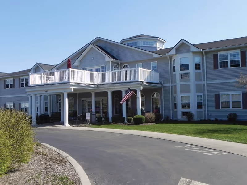 Exterior front view of a two-story senior living facility building with gray siding, white trim, and a covered entrance supported by white columns. There is a balcony with white railing above the entrance, an American flag hanging near the entrance, and a paved driveway leading up to the building. The sky is clear and blue.