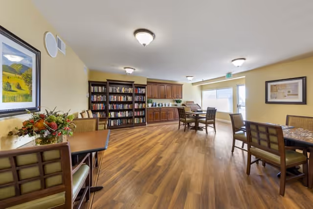 Warm lit senior living common area with wood floors, bookshelves, and multiple tables with chairs.