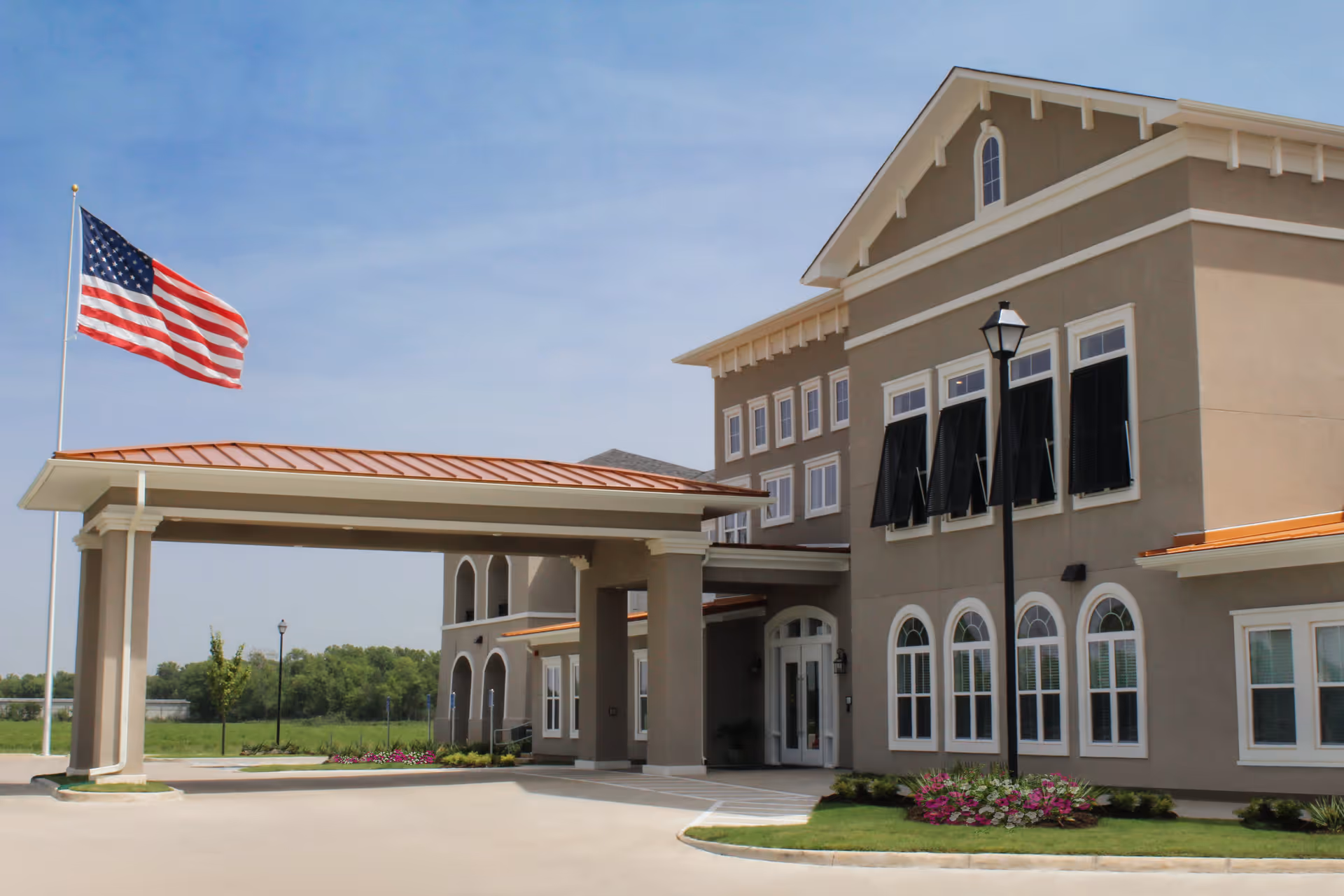 Exterior view of The Blake at Bossier City senior living facility showing the entrance with a covered driveway, beige building with multiple windows, an American flag on a flagpole, and landscaped greenery with flowers.