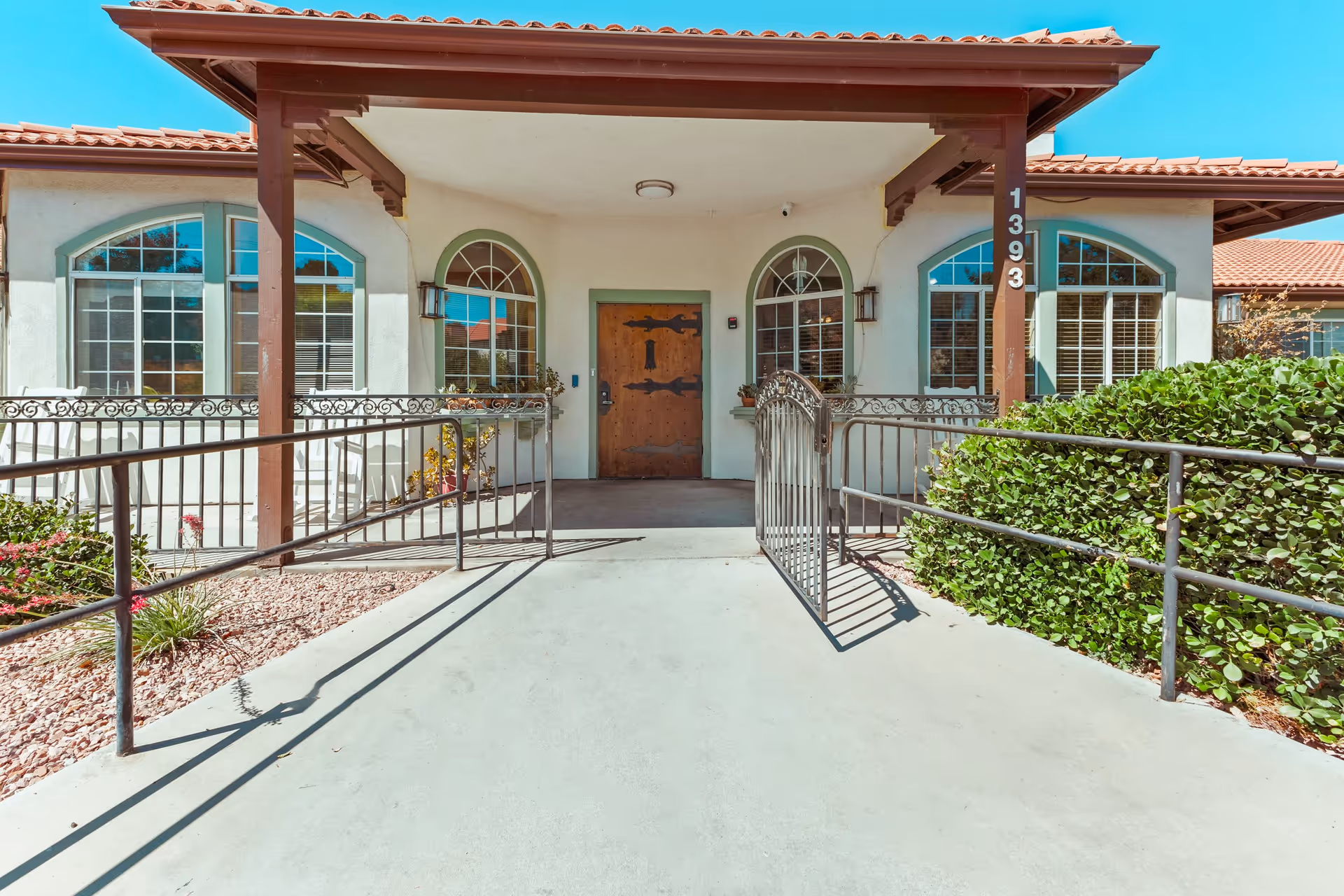 Front entrance of Buena Vista Assisted Living facility featuring a covered porch with a wooden door, arched windows on either side, a metal gate, and a ramp with railings leading up to the door. The building has a tiled roof and is surrounded by bushes and landscaping with rocks.