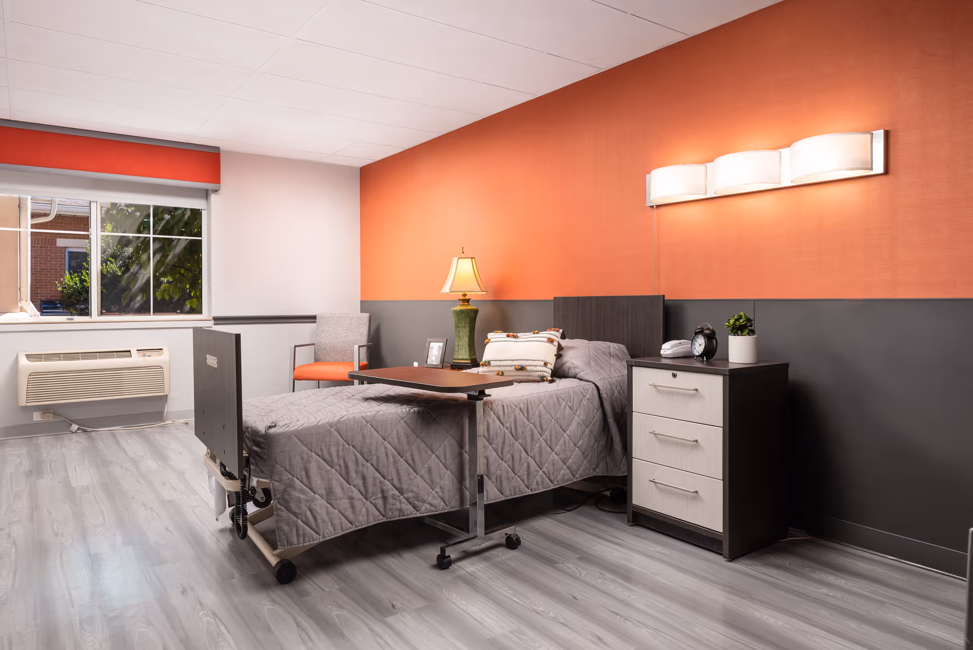 Senior living bedroom with a bed, nightstand, lamp, and orange-accent wall.