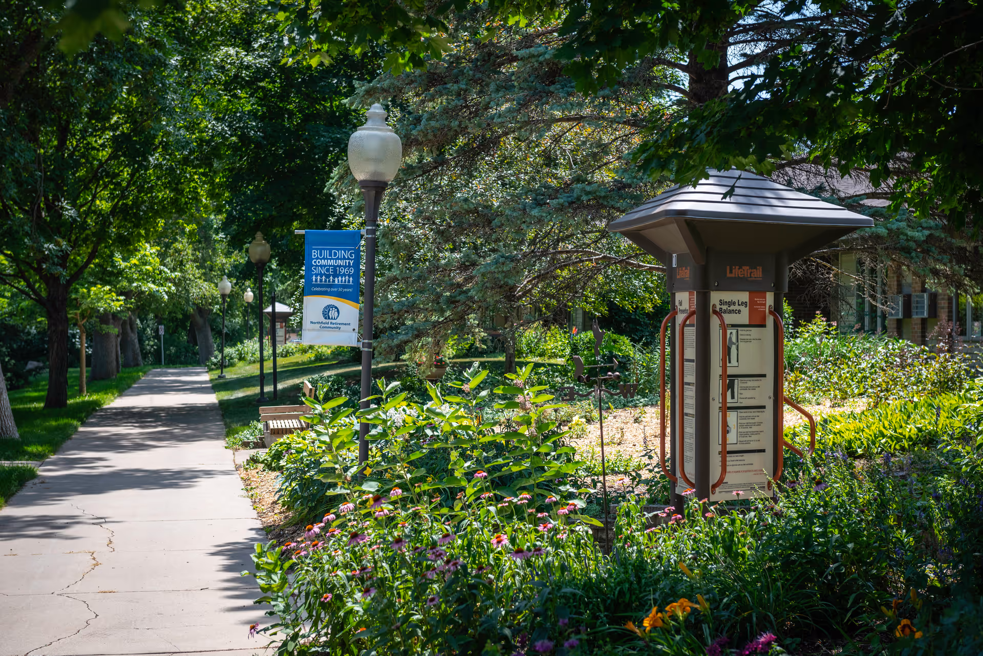 A sunny outdoor pathway lined with trees, lampposts, and lush greenery at Northfield Retirement Community by Vivie. There is a blue sign on a lamppost that reads 'Building Community Since 1969' and an exercise station with instructions for balance exercises surrounded by flowers and plants.
