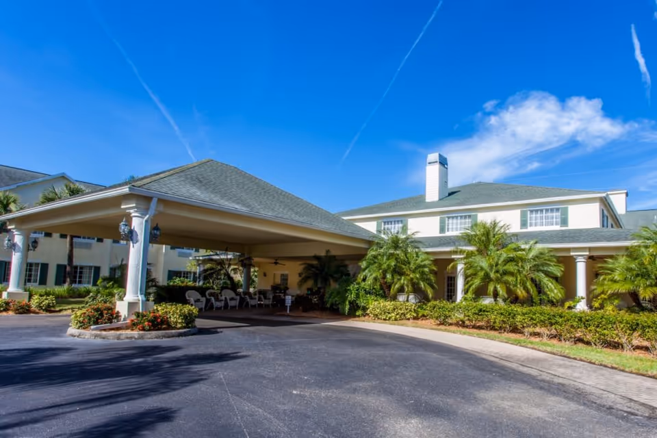 Exterior view of Lexington Manor senior living facility showing a covered entrance with white columns, surrounded by palm trees and landscaping under a clear blue sky.
