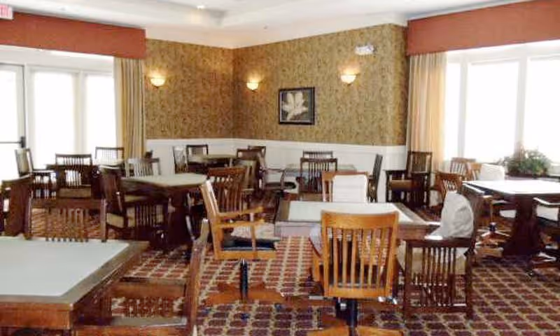 A dining room with multiple wooden tables and chairs arranged neatly on a patterned carpet. The room has large windows with beige curtains allowing natural light to enter. The walls are decorated with wallpaper and a framed picture, and there are wall-mounted lights providing additional illumination.