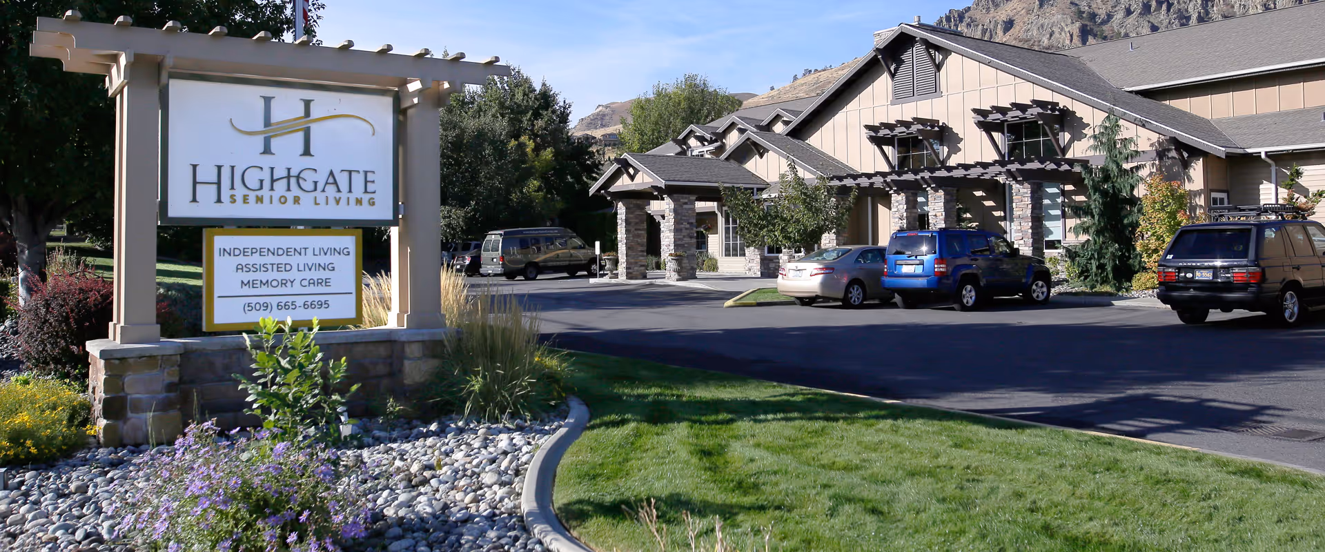 Exterior view of Highgate Senior Living facility in Wenatchee, showing the building entrance with parked cars and a large sign displaying the facility name and contact information, surrounded by landscaping with grass, flowers, and trees.
