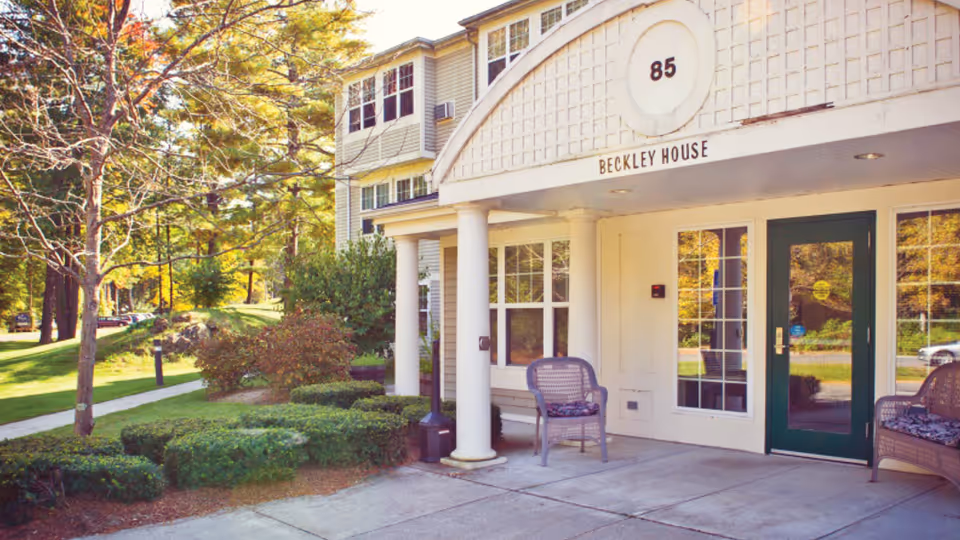 Front exterior of a senior living building with pillars, a covered entrance, and chairs.